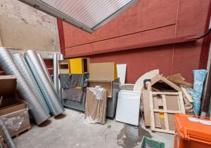 Une cour intérieure en béton encombrée de débris de construction, de conduits métalliques flexibles, de meubles cassés, de palettes en bois et de déchets mélangés devant un mur rouge