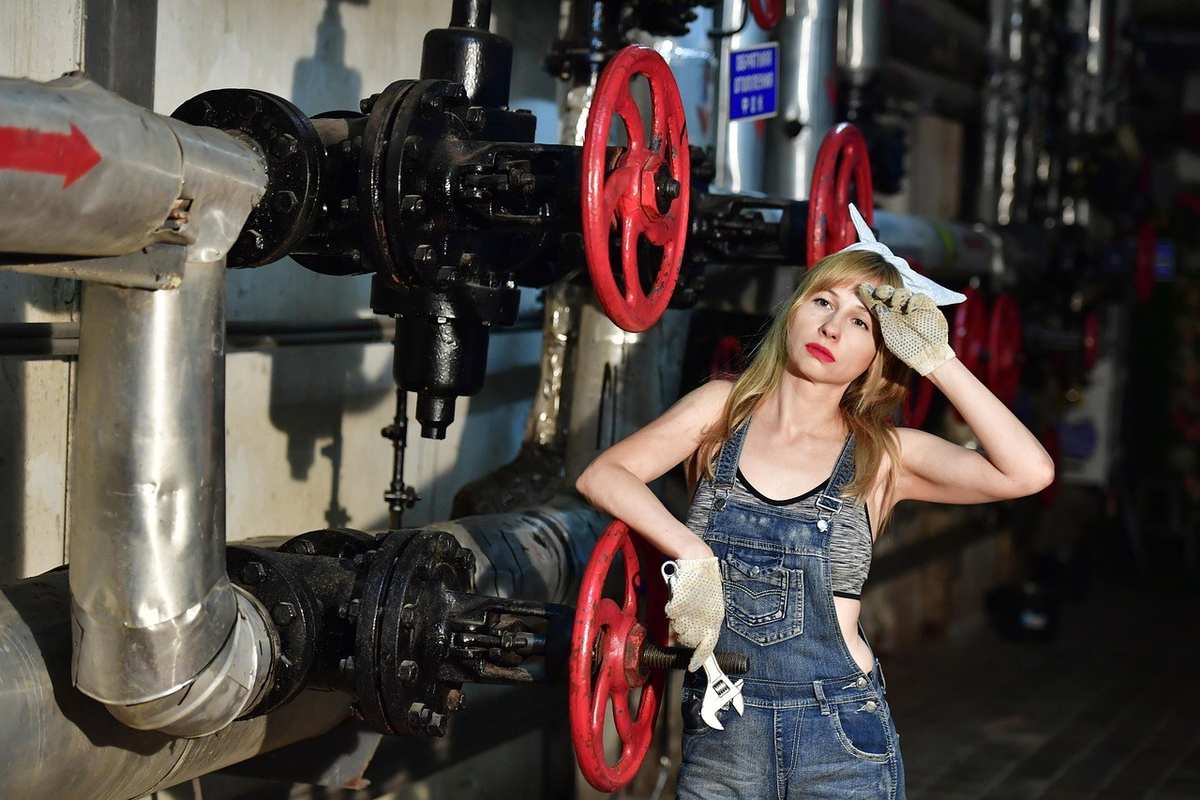 Femme en overalls devant tuyaux et manivelles rouges, pose contemplative