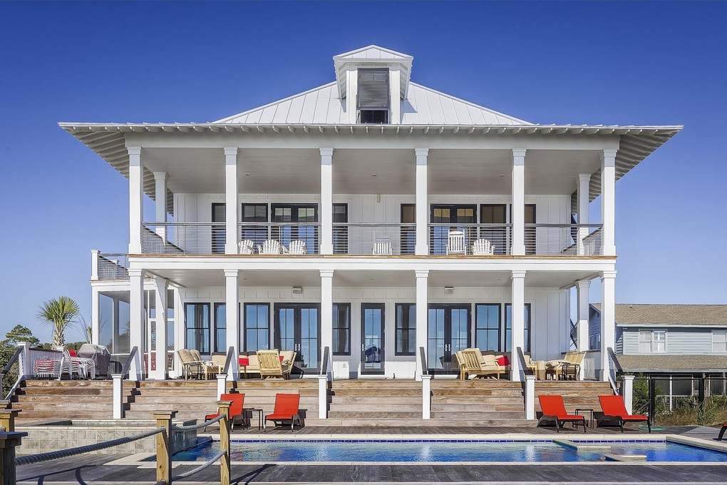 Maison blanche à deux étages avec balcon, piscine, chaises rouges, ciel bleu, végétation